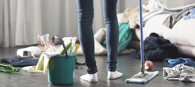 Mujer preparada para comenzar la limpieza del hogar, con productos de limpieza y un trapeador en mano, mientras un entorno desordenado se observa de fondo.